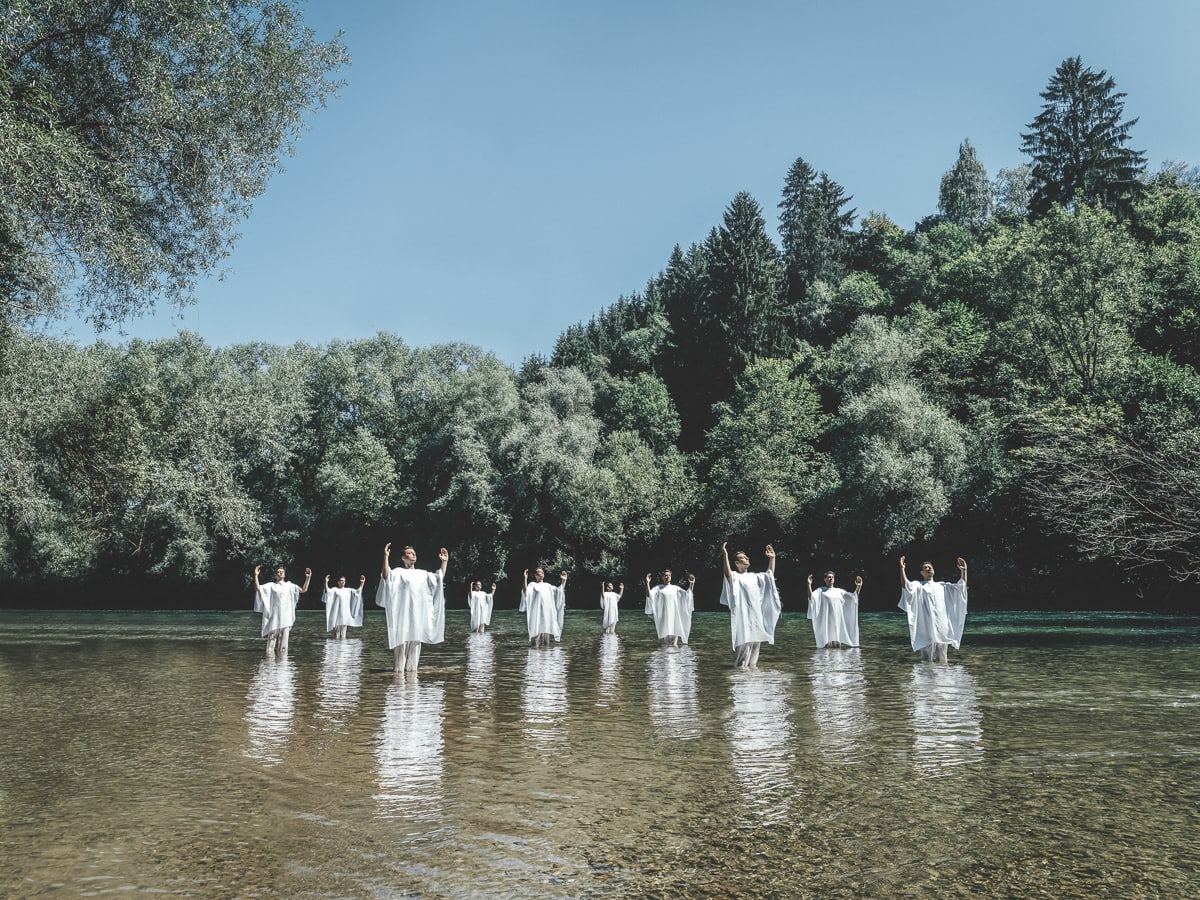 Ein flacher See mit Kiesgrund, den man sieht. Blauer Himmel, um den See Bäume. im See stehen zehn weiß gekleidete Personen (Männer), alle mit erhobenen Armen. Titel des Bildes: 'Light Workers and Light Pioneers'. Foto: Klaus Pichler