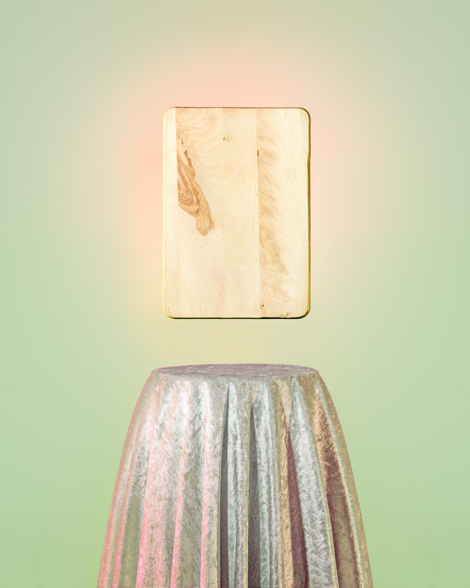 "harmonie engergie transformer" - ein Schneidebrett. Foto: Klaus Pichler Bild mit lindgrünem Hintergrund. Unten ein runder Stehtisch mit silbrig glänzendem Überwurf, an der Wand in rosa Corona der "harmonie engergie transformer" - ein Schneidebrett. Foto: Klaus Pichler