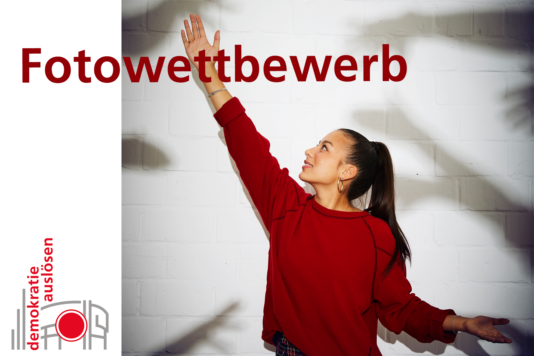 Eine junge Frau steckt den rechten Arm zum Himmel, auf der Wand hinter ihr mehrere Schattenhände. Dazu der Schriftzug "Fotowettbewerb" und das Logo zum Wettbewerb "Demokratie auslösen" 