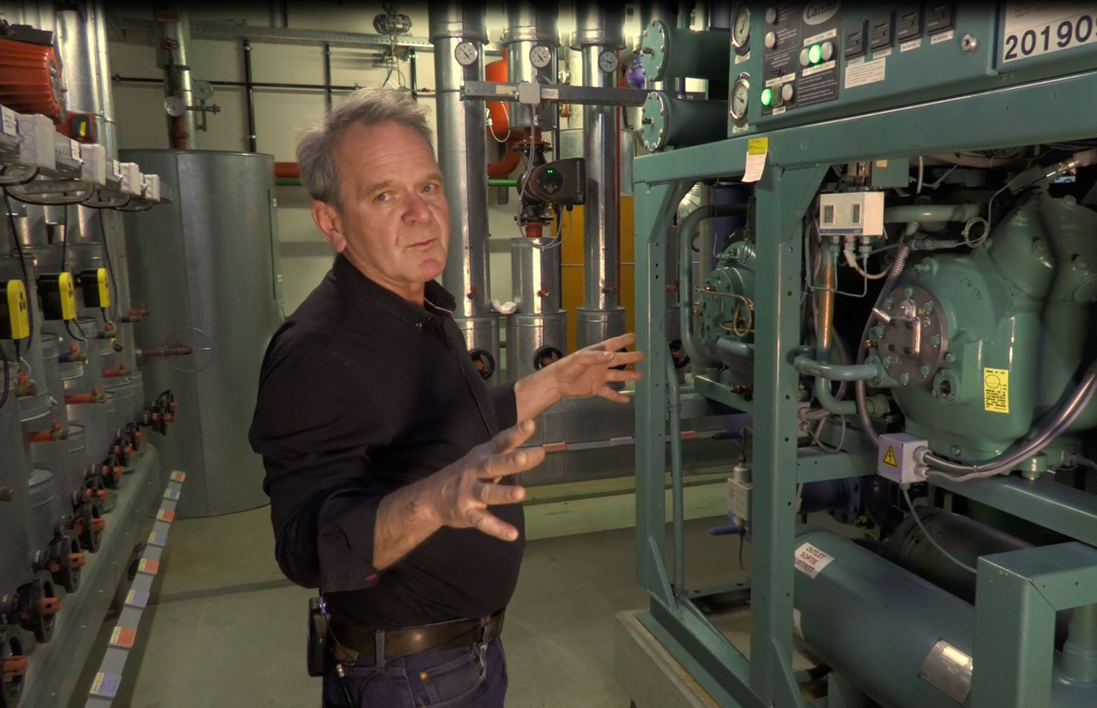 In der Lüftungs- und Kühlungsanlage des Stadthauses. Teil der Führung durch die technischen Betriebsräume mit dem Technischen Leiter des Stadthauses Paul Stauber. Filmstill Der technische Leiter des Stadthauses im schwarzen Hemd steht vor der grünen Kältemaschine, die das Stadthaus temperiert.