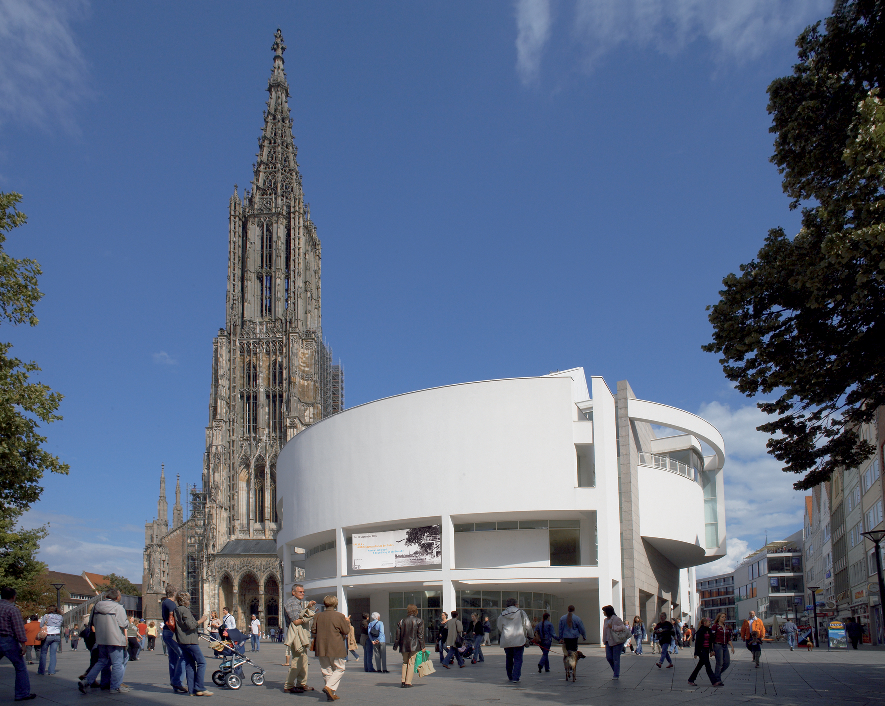 Das Stadthaus und das Ulmer Münster. Foto: Nik Schölzel Das Stadthaus und das Ulmer Münster: im Vordergrund der weiße Baukörper des Stadthauses, links dahinter ragt der Münsterturm auf. Im Vordergrund laufen zahlreiche Menschen.