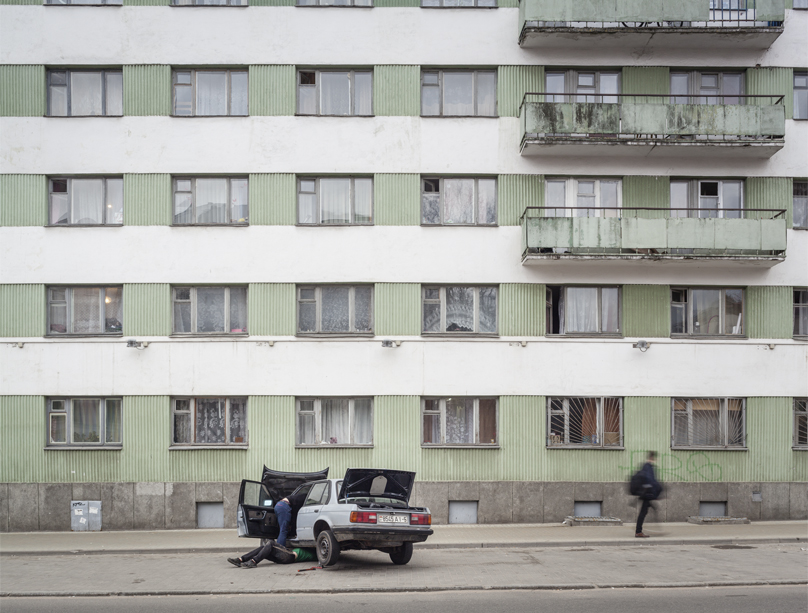 Peter Bialobrzeski: Minsk Diary. Vor einem tristen, kahlen Häuserblock liegt eine Person unter einem weißen Pkw, Kofferraum, Kühlerhaube und Fahrertür sind offen. Eine weiter Passant läuft am Haus vorbei.